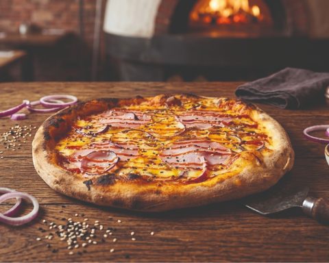 Pizza garnie de viande et d'oignons rouges, posée sur une table en bois près d'un four.
